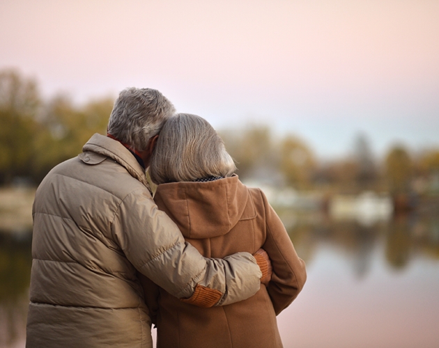 An older man wraps his arm around his wife while they look out over a lake.