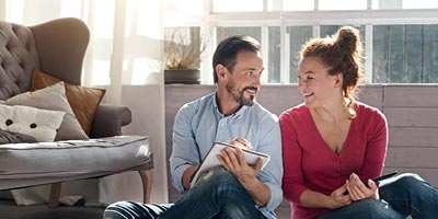 A young couple sits on the floor, smiling and looking at a notebook and iPad, as if creating a household budget. 