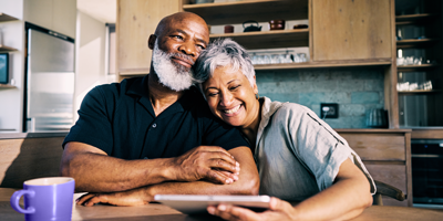 Older African American couple sitting arm-in-arm looking at tablet in their kitchen.
